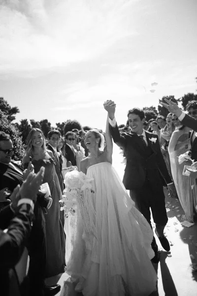 Ceremony exit with a flower petal toss as the bride and groom walk hand in hand, smiling amid cheering guests along the tree-lined walkway.