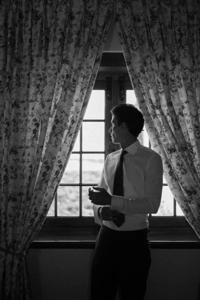 Groom getting ready by the window, straightening his necktie beside floral curtains with trees visible outside.