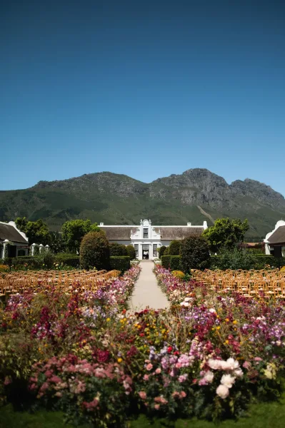 Ceremony setup with outdoor ceremony decor and bentwood chairs on a lawn before a manor house, framed by garden hedges and a mountain range.