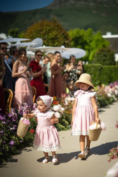 Flower girl processional with children walking down the aisle, carrying woven baskets in a garden setting beneath a mountain and blue sky.