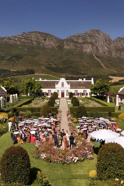 Ceremony moment at a garden wedding ceremony as the couple exchanges vows beside blush aisle florals, mountains and a manor house beyond.