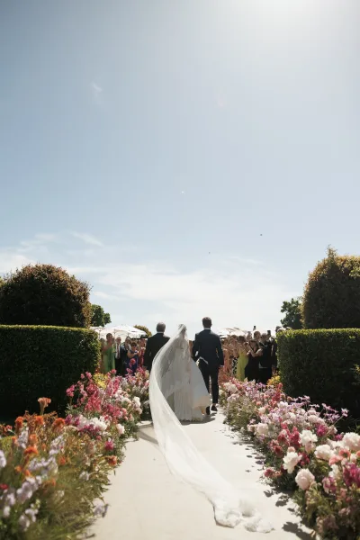 Wedding processional with a bride walking down the aisle along a paved garden path under a blue sky, her bridal veil softly trailing behind.