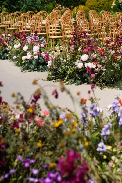 Ceremony aisle design for a garden ceremony setup, grounded arrangements of peonies lining a grass lawn path amid shrubs and trees.