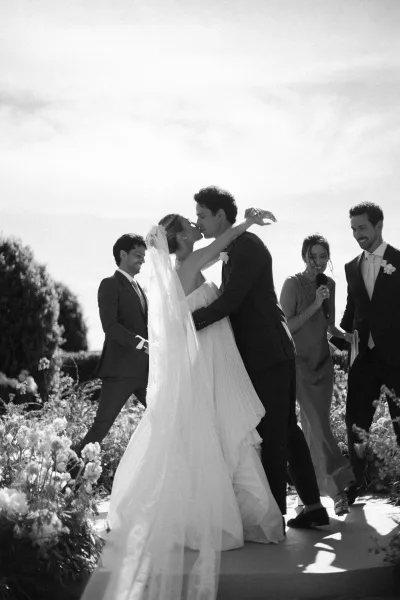 Wedding kiss moment and tender ceremony moment as the bride and groom embrace in a sunlit garden, her veil draped back against green hedges.