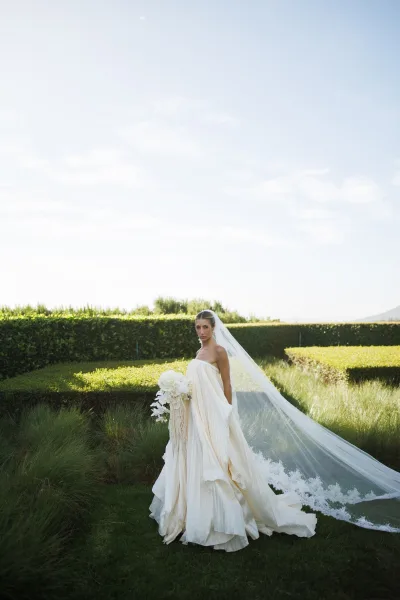 Bridal portrait of a bride holding a bouquet, her long veil flowing beside garden hedges on a sunlit lawn under a clear blue sky.