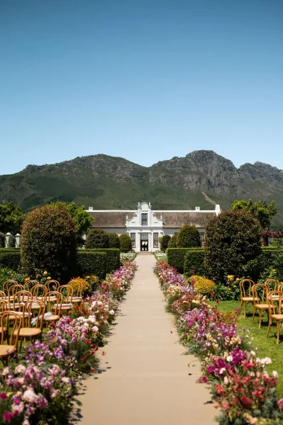 Ceremony aisle design for garden ceremony decor, flanked by bentwood chairs, leads across a manicured lawn toward a manor with mountain views.