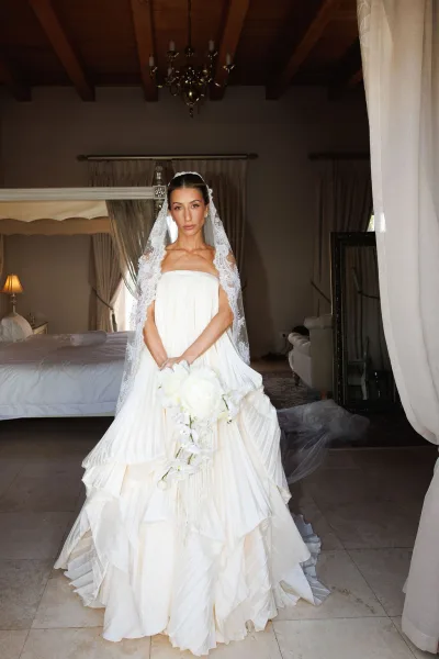 Bridal portrait of a bride holding a bouquet with a long lace veil trailing, in a bedroom with a canopy bed and chandelier.