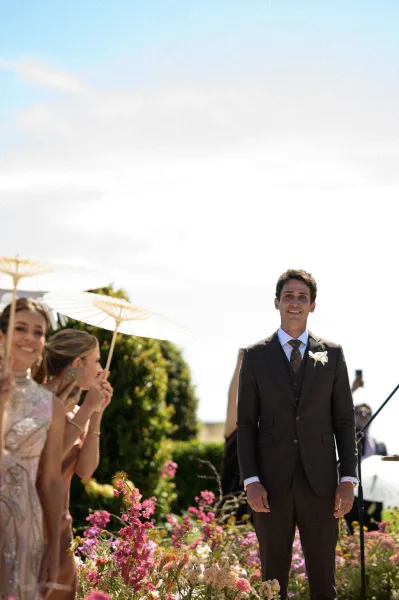 Ceremony moment with the groom at altar, his white boutonniere bright against his lapel, on a sunlit lawn framed by blue sky and garden hedges.