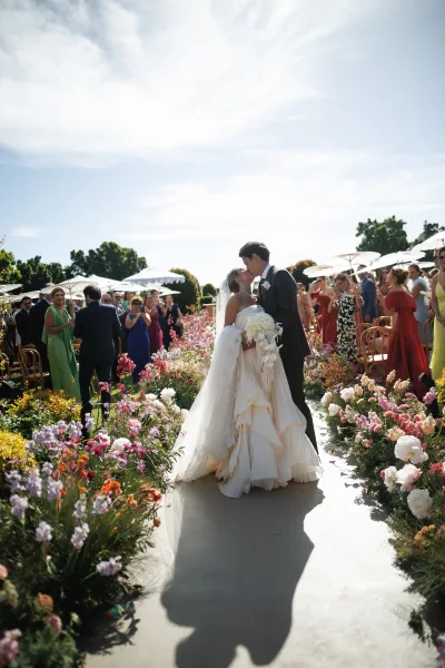 Wedding kiss portrait as the couple share a ceremony exit kiss along a garden aisle, guests with parasols cheering under blue sky and trees
