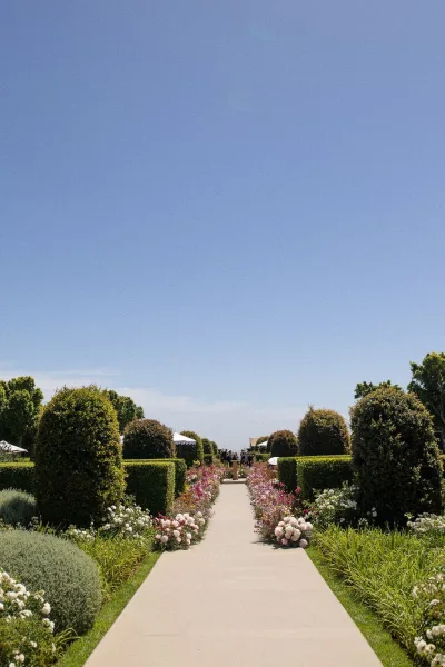 Ceremony aisle design with wedding aisle flowers in blush and white edging a long path, flanked by topiary, under a blue sky amid garden trees