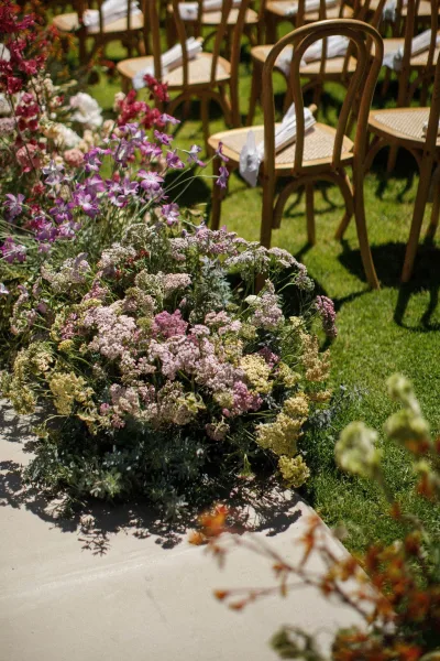 ceremony aisle flowers create a grounded floral aisle beside wooden chairs on a paved aisle across the grass lawn