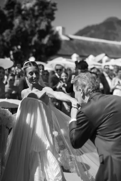 Ceremony moment with a wedding hand kiss as a partner bows to kiss the bride's hand, her veil trailing, against mountain trees and distant rooftops.
