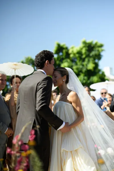 Ceremony moment as a wedding forehead kiss is shared between bride and groom, guests shaded by parasols at a sunny garden ceremony.