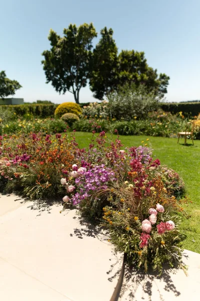 Ceremony aisle flowers anchor lush outdoor ceremony decor, blooming with peonies along a lawn path framed by garden trees and hedges.