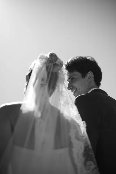 Couple portrait of a bride with veil facing her groom, lace trim detail framing her shoulders as they share a tender, romantic moment.