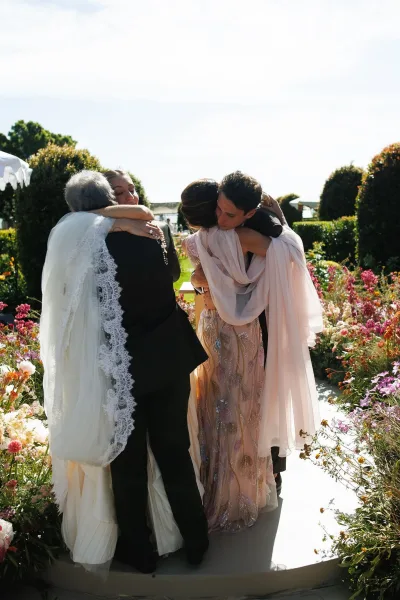 Ceremony moment capturing a family embrace at ceremony as the bride hugs a parent, her beaded gown visible in a garden by hedges and trees.