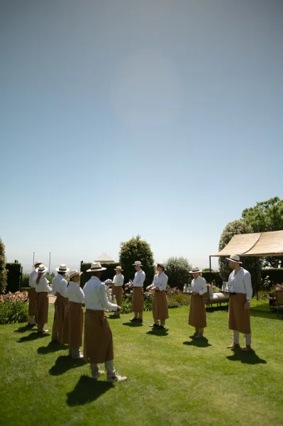 Wedding servers carrying cocktail hour drinks in straw hats on a garden lawn beneath a white canopy tent under a blue sky.