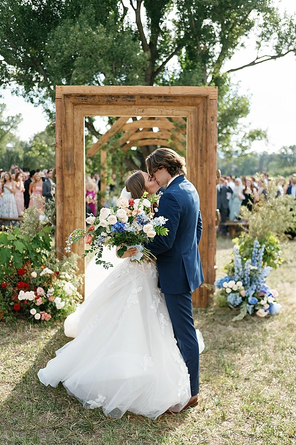 Wedding kiss portrait of bride and groom kissing beneath a rustic wooden arch with blue and white bouquet, guests on a tree-lined lawn
