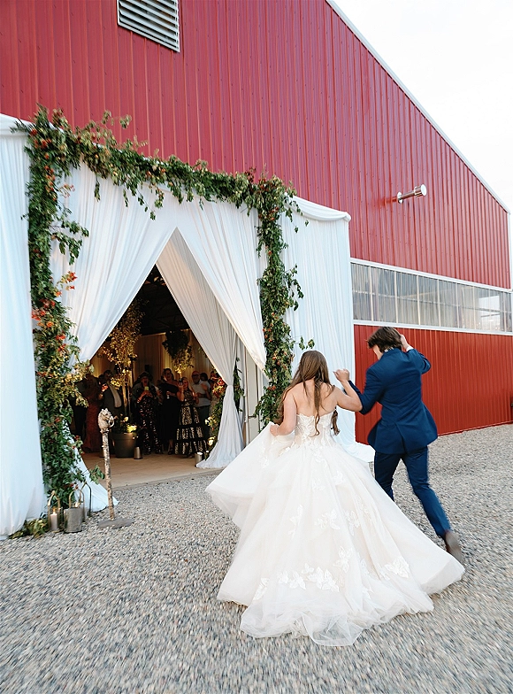 Wedding grand entrance as newlyweds enter the reception through a draped greenery doorway, bride holding her train beside groom, red barn behind