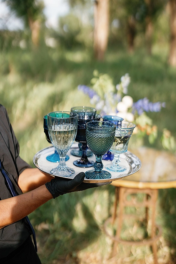 Wedding drinkware with colored glass goblets and champagne flutes on a serving tray, held by black-gloved hands on a wooden table outdoors