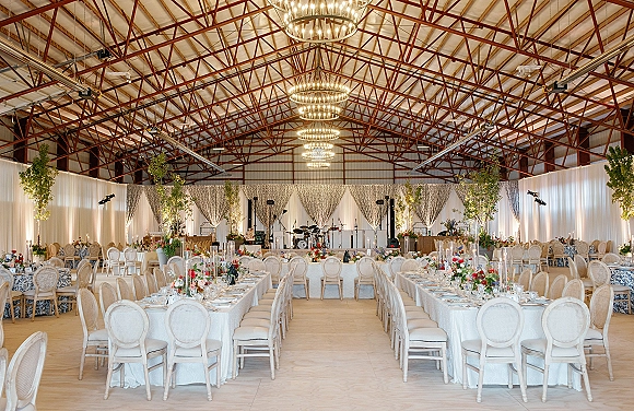 Reception tablescape with wedding reception layout of long banquet tables in white linen, floral centerpieces, taper candles, chandeliers in a warehouse hall