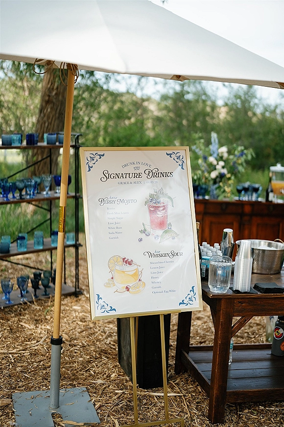 Signature drinks sign on an easel with illustrated cocktail menu, framed in gold beside dispensers and glassware at an outdoor bar setup