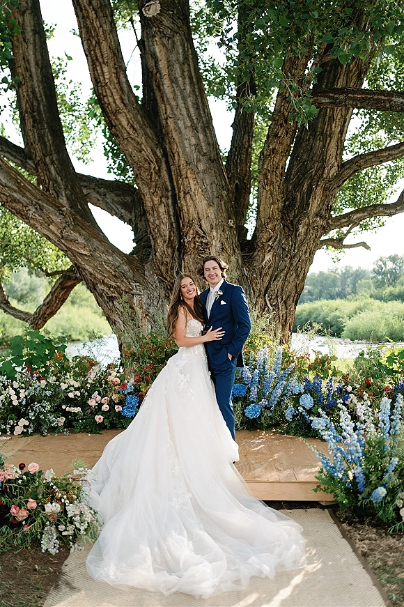 Couple portrait of bride in strapless lace gown with long train and groom in navy suit under a large tree by the river, floral meadow behind