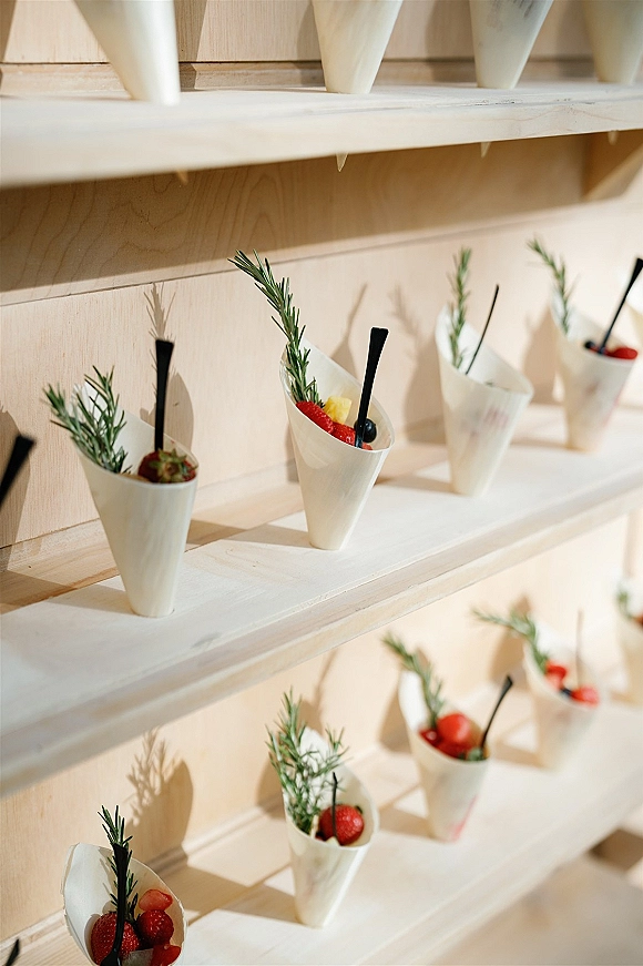 Wedding dessert display of berry dessert cups in paper cones with fresh berries and rosemary sprigs on wooden shelves against a light wood wall