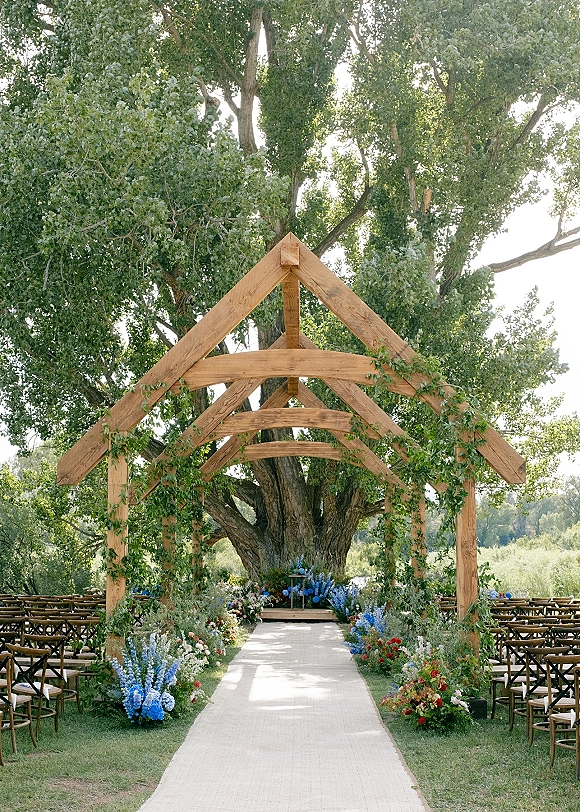 Ceremony aisle decor with an outdoor wedding ceremony setup featuring a white runner, crossback chairs, and wildflowers under a large tree