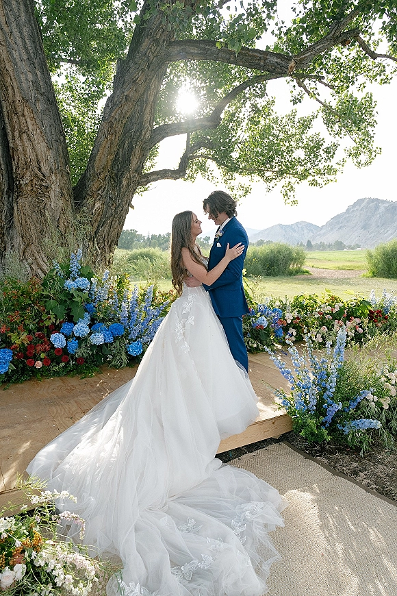 Couple portrait of bride and groom embrace beneath a large tree, her lace train flowing beside blue hydrangea with sun flare and mountains