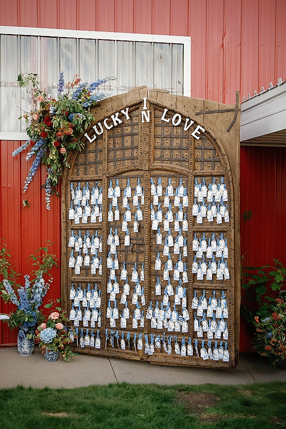Wedding escort display on wood arched doors with place cards tied by blue ribbons, framed by roses and delphinium against a red barn wall