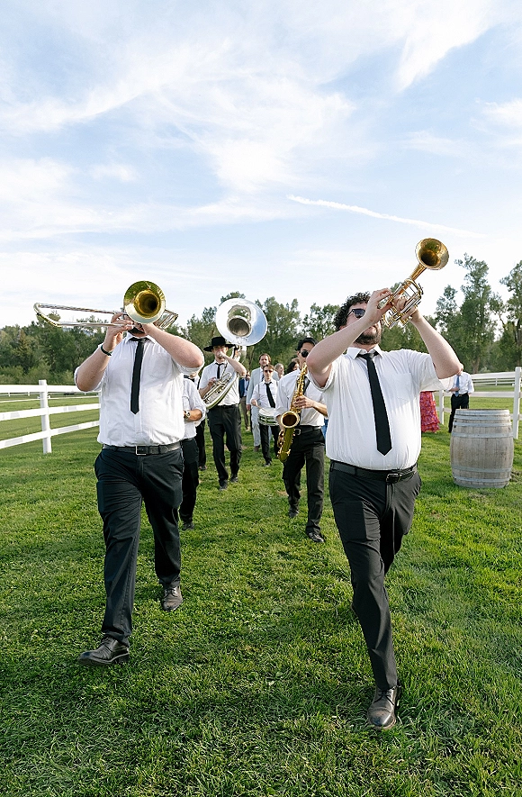 Wedding band in white shirts and black ties marching across a grassy lawn, playing brass instruments by a white fence and trees