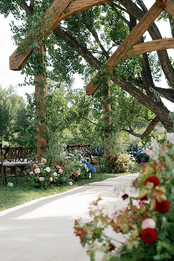 Ceremony setup for an outdoor wedding ceremony with a wood wedding arch, greenery garland, floral aisle runner, and chairs beneath trees
