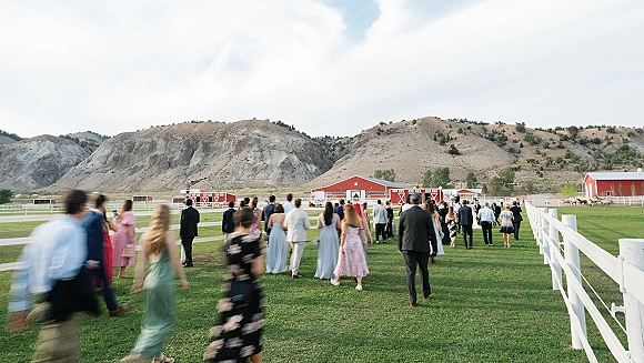 Wedding guests walking in formal attire across a green lawn toward a red barn, with a white fence and mountains under cloudy sky