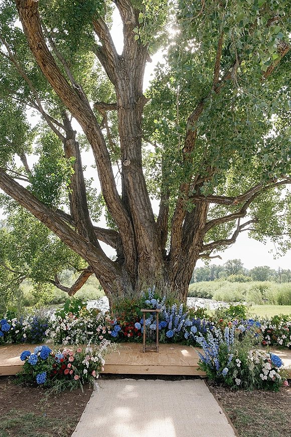 Outdoor ceremony altar with tree wedding altar florals in hydrangeas and roses on a wood platform with rug runner by a river under canopy