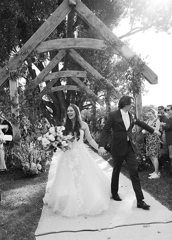 Wedding recessional as bride and groom walking down aisle holding hands, bouquet in hand, under a greenery-wrapped wooden arbor with guests cheering