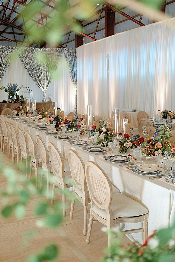 Reception tablescape with a long banquet table wedding setup, patterned plates, florals, taper candles in glass cylinders, in a draped barn interior