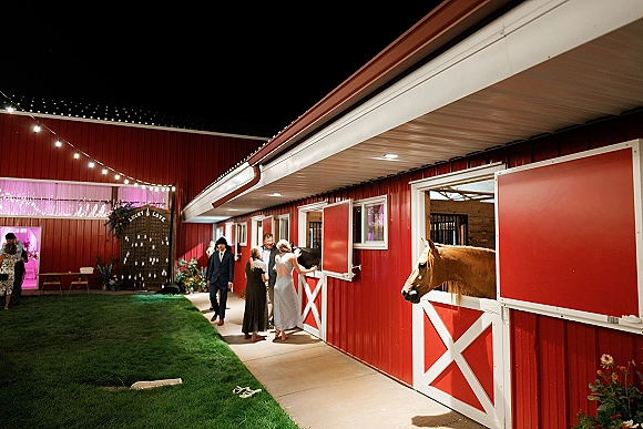 Barn wedding reception entrance with string lights, floral escort card display, and open barn doors on a grassy lawn at night