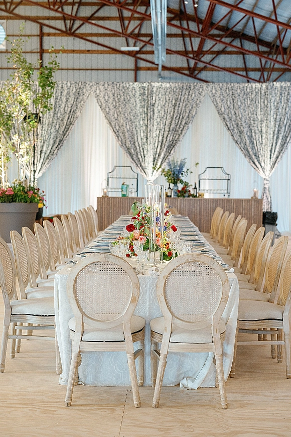 Reception tablescape on a long banquet table wedding setup with white tablecloth, cane back chairs, florals, candles, and barn trusses backdrop