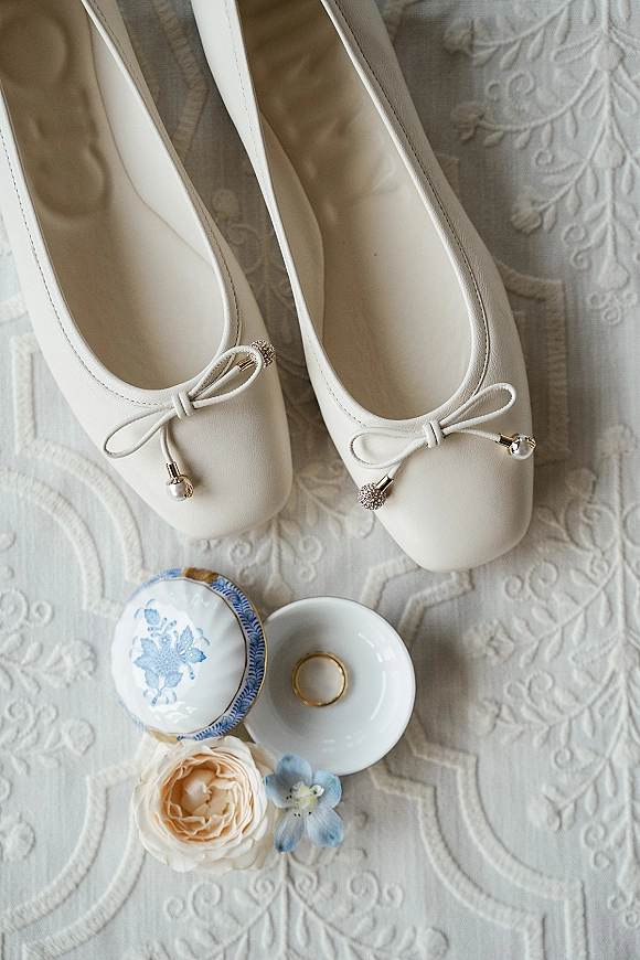 Bridal shoes flatlay of white bridal flats with pearl bow detail, gold ring and dish, and blue trinket box on embroidered lace background