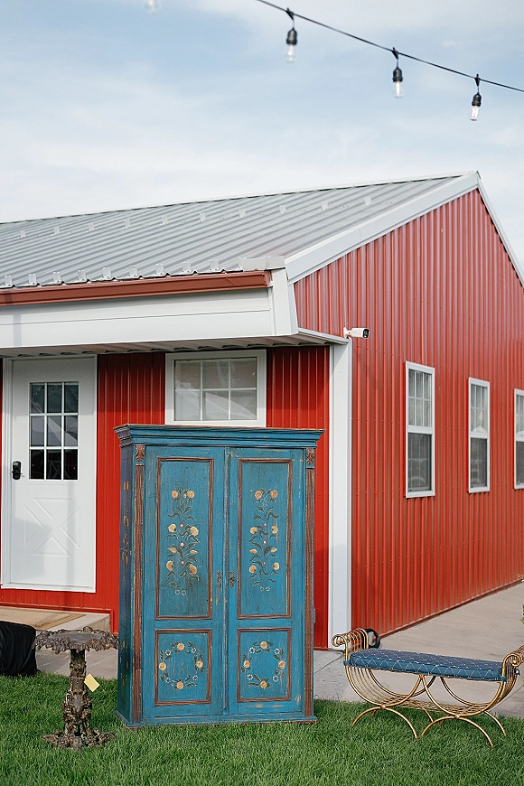 Wedding lounge area outdoor wedding lounge with string lights, vintage blue cabinet, and upholstered bench set by a red barn on the lawn