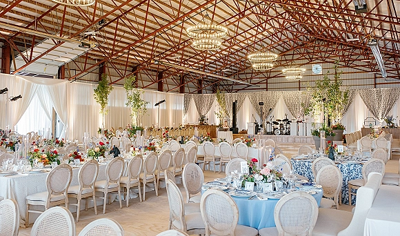 Reception tablescape with wedding reception decor in blue and white linens, cane-back chairs, floral centerpieces, candles, and chandeliers in an industrial hall
