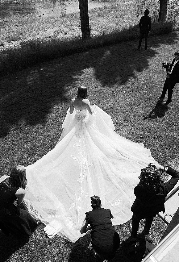 Bride with train in a cathedral train wedding dress as bridesmaids arrange her long tulle lace gown on a sunlit lawn by trees and pond