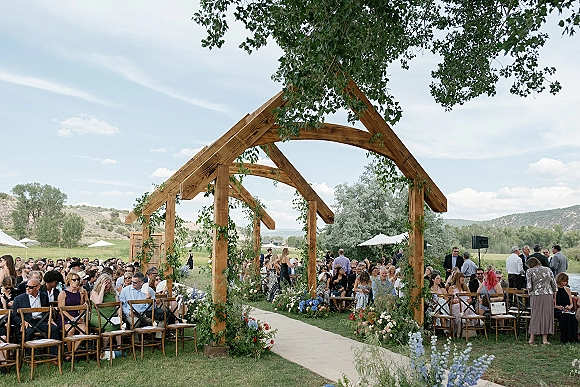 Ceremony setup for an outdoor wedding ceremony with wooden arbor and greenery garlands, aisle runner and ground florals on lawn with mountains