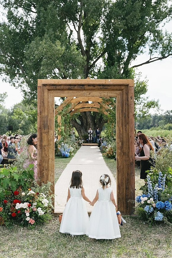 Flower girl processional of two flower girls holding hands in white dresses, carrying a woven basket down an aisle runner to a wooden arch outdoors