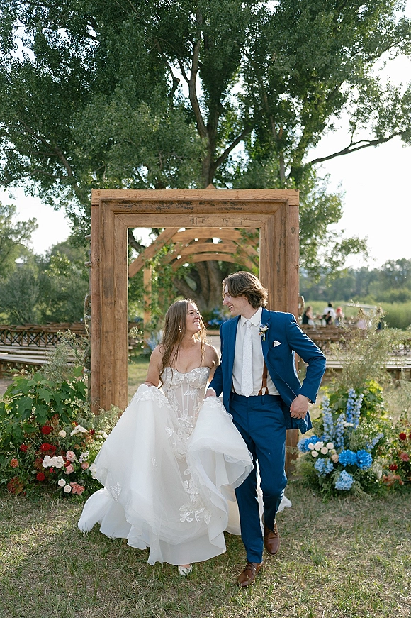 Couple portrait of bride and groom walking down the garden wedding aisle, her lace dress held up, his blue suit, smiling under a wooden arch