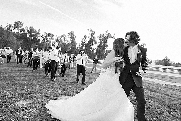 Wedding kiss portrait of bride and groom kissing while walking with drinks, bride in ball gown, with brass band and guests in a grassy field