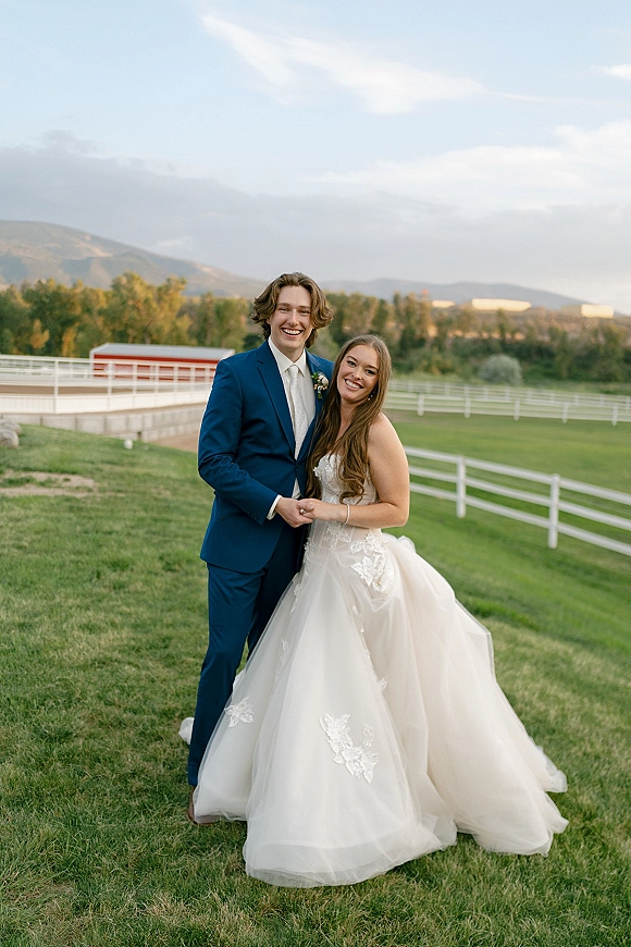 Couple portrait of bride in lace tulle gown leaning on groom in blue suit, holding hands in grassy field by barn and mountains