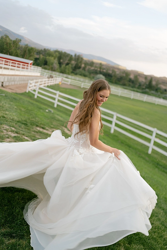 Bridal portrait of a bride twirling her tulle gown, looking over her shoulder on a green lawn with a white fence and barn backdrop