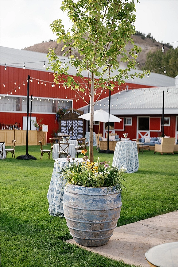 Outdoor reception decor with backyard wedding reception cocktail tables under string lights, lounge seating and bar by a red barn with mountains behind